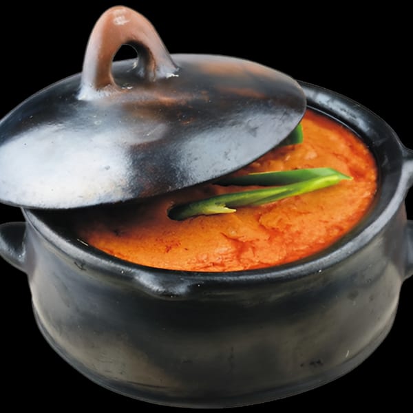 A black clay pot with a slightly ajar lid, revealing vibrant orange Ethiopian stew garnished with green onions.